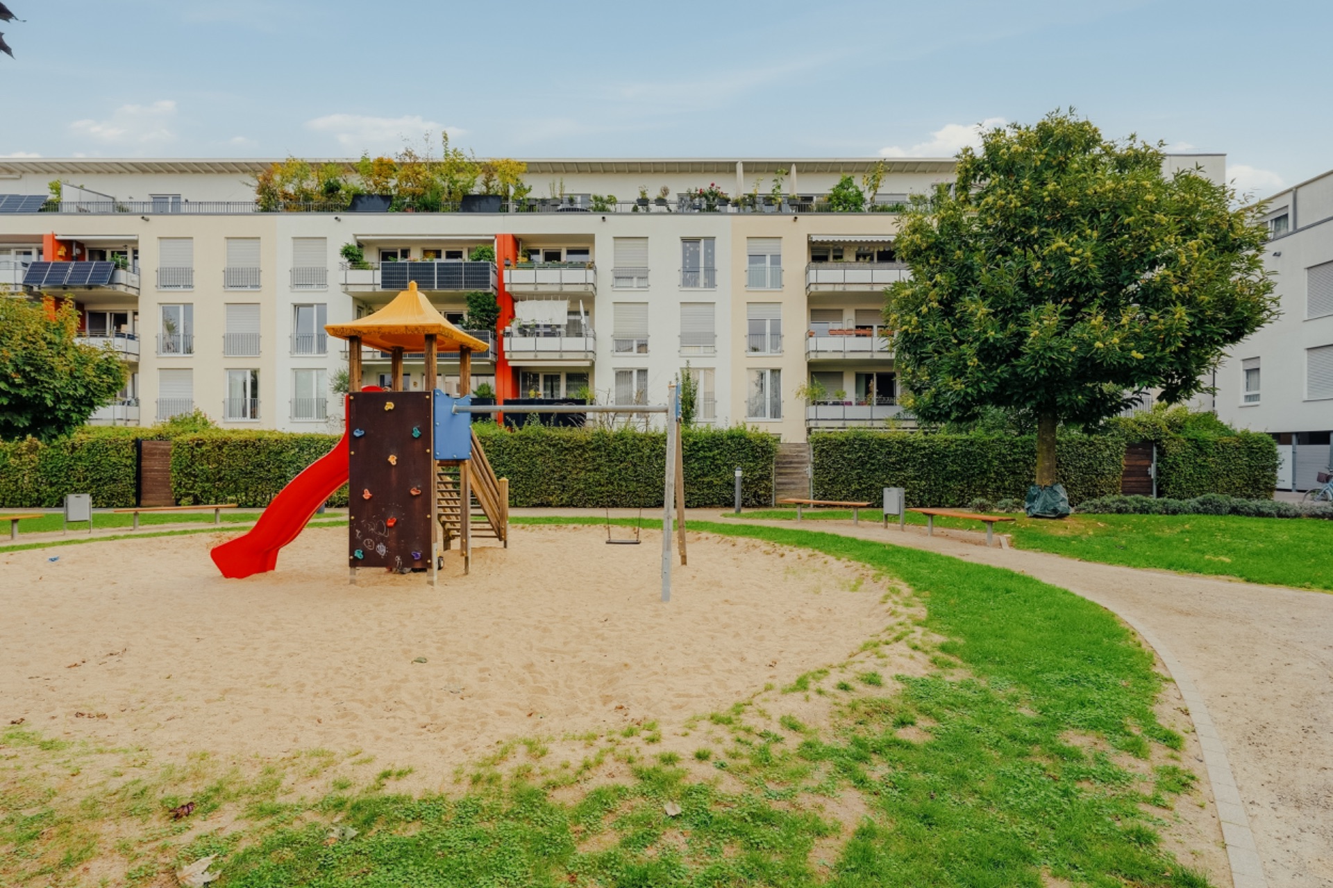 Spielplatz mit Rutsche und Kletterwand vor einem Mehrfamilienhaus mit Balkonen.