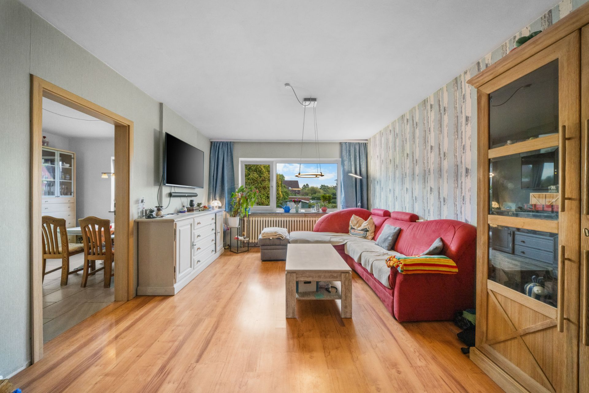 Wohnzimmer mit rotem Sofa, Couchtisch, Holzschrank und Blick auf großes Fenster, Esszimmer im Hintergrund.