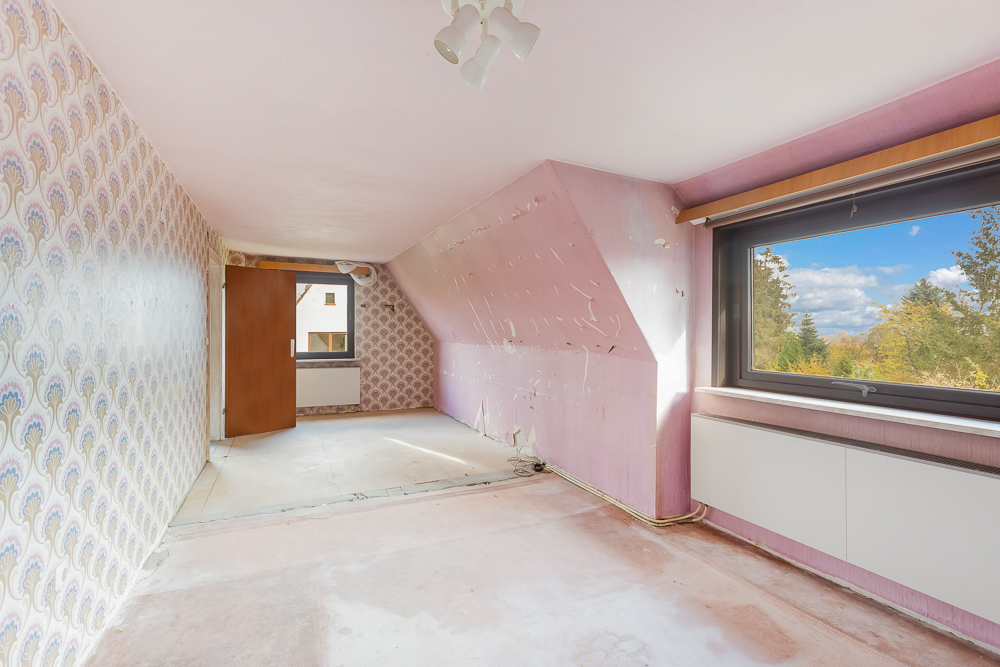 Leeres Zimmer mit Tapeten, schräger Decke, großem Fenster und Laminatboden.