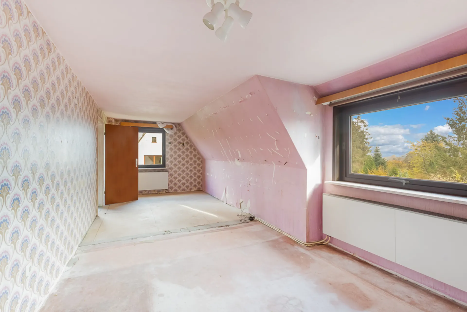 Leeres Zimmer mit Tapeten, schräger Decke, großem Fenster und Laminatboden.