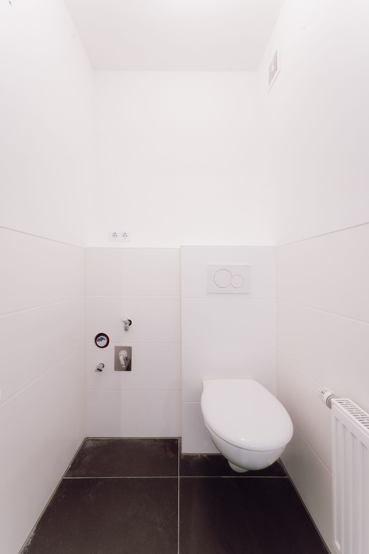 Weiß gefliestes WC mit Wandtoilette, dunklem Boden und Heizung rechts.