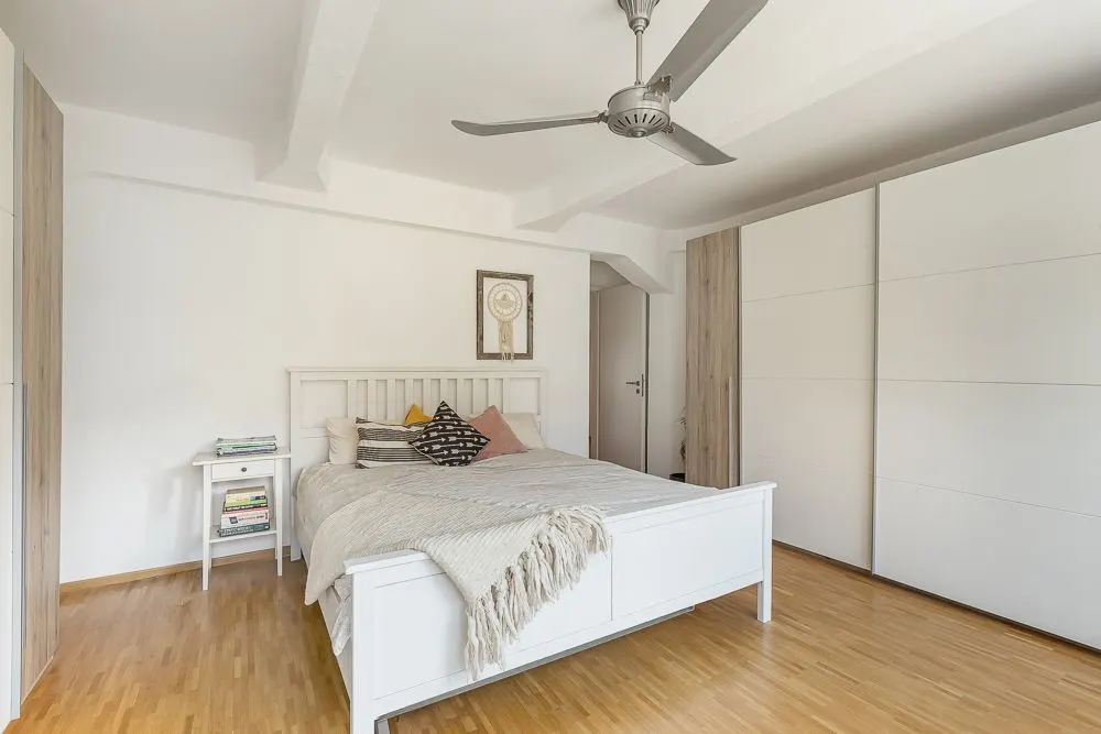 Helles Schlafzimmer mit weißem Bett, Nachttisch, Deckenventilator und großem Kleiderschrank.