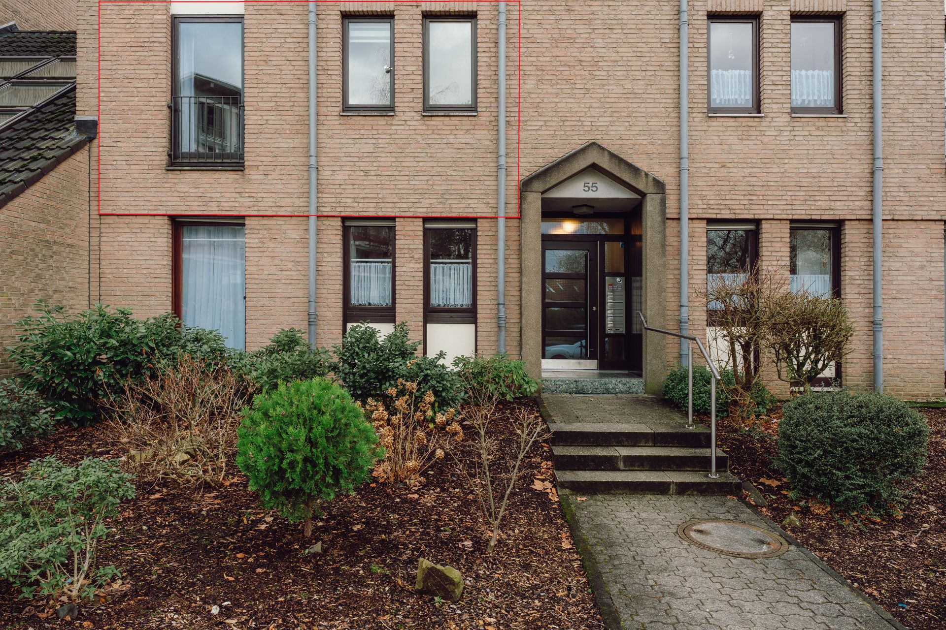 Eingang eines Mehrfamilienhauses mit Stufen, Sträuchern und Büschen im Vordergrund.