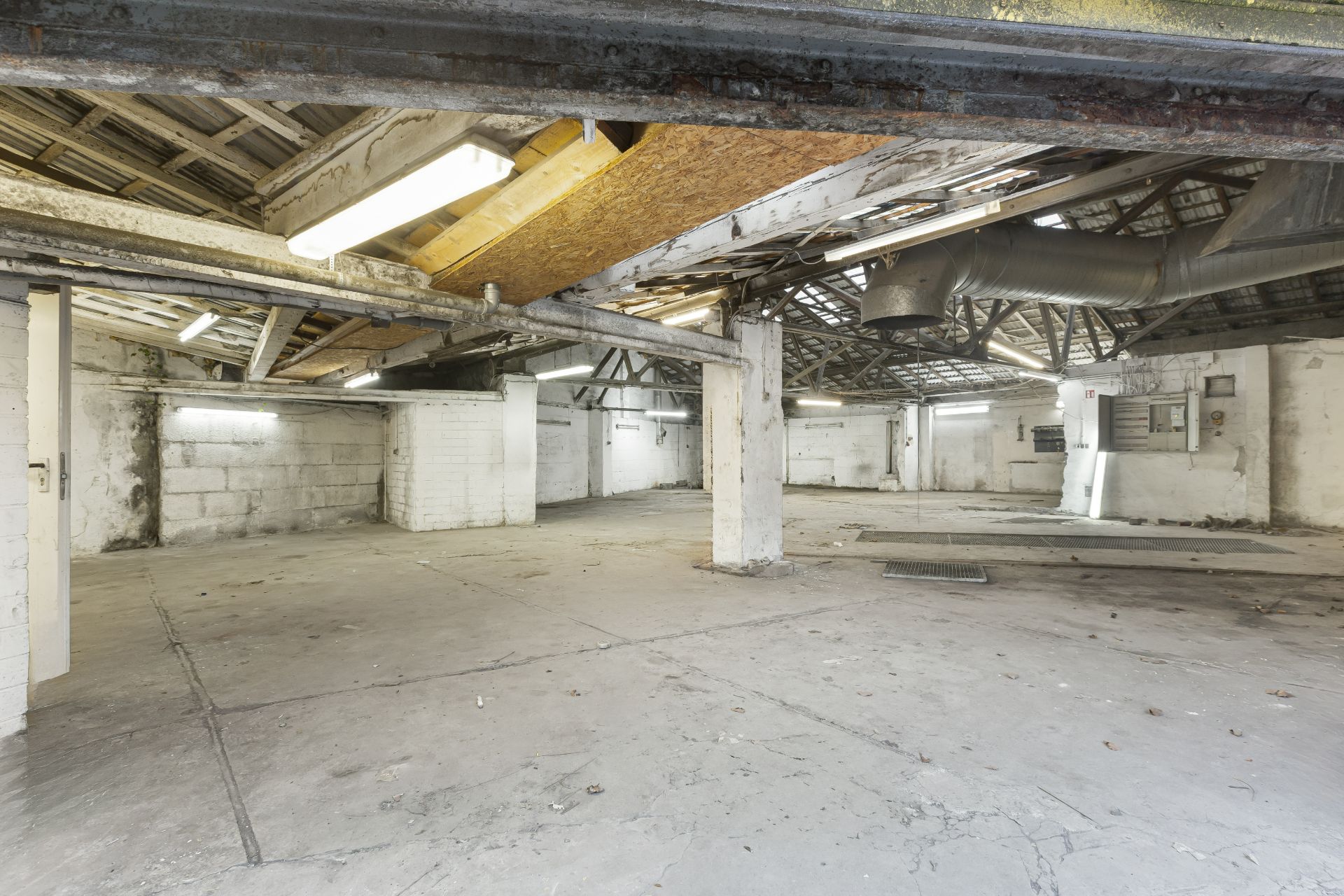 Leere, große Halle mit Betonboden, weißen Wänden und sichtbaren Rohren an der Decke.