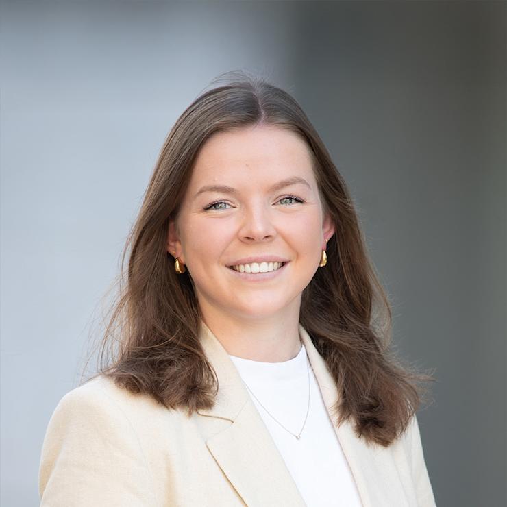 Junge Frau mit langen braunen Haaren im dunkelblauen Blazer steht vor einer Glaswand.