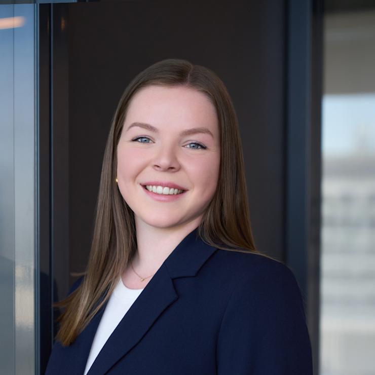 Junge Frau mit langen braunen Haaren im dunkelblauen Blazer steht vor einer Glaswand.