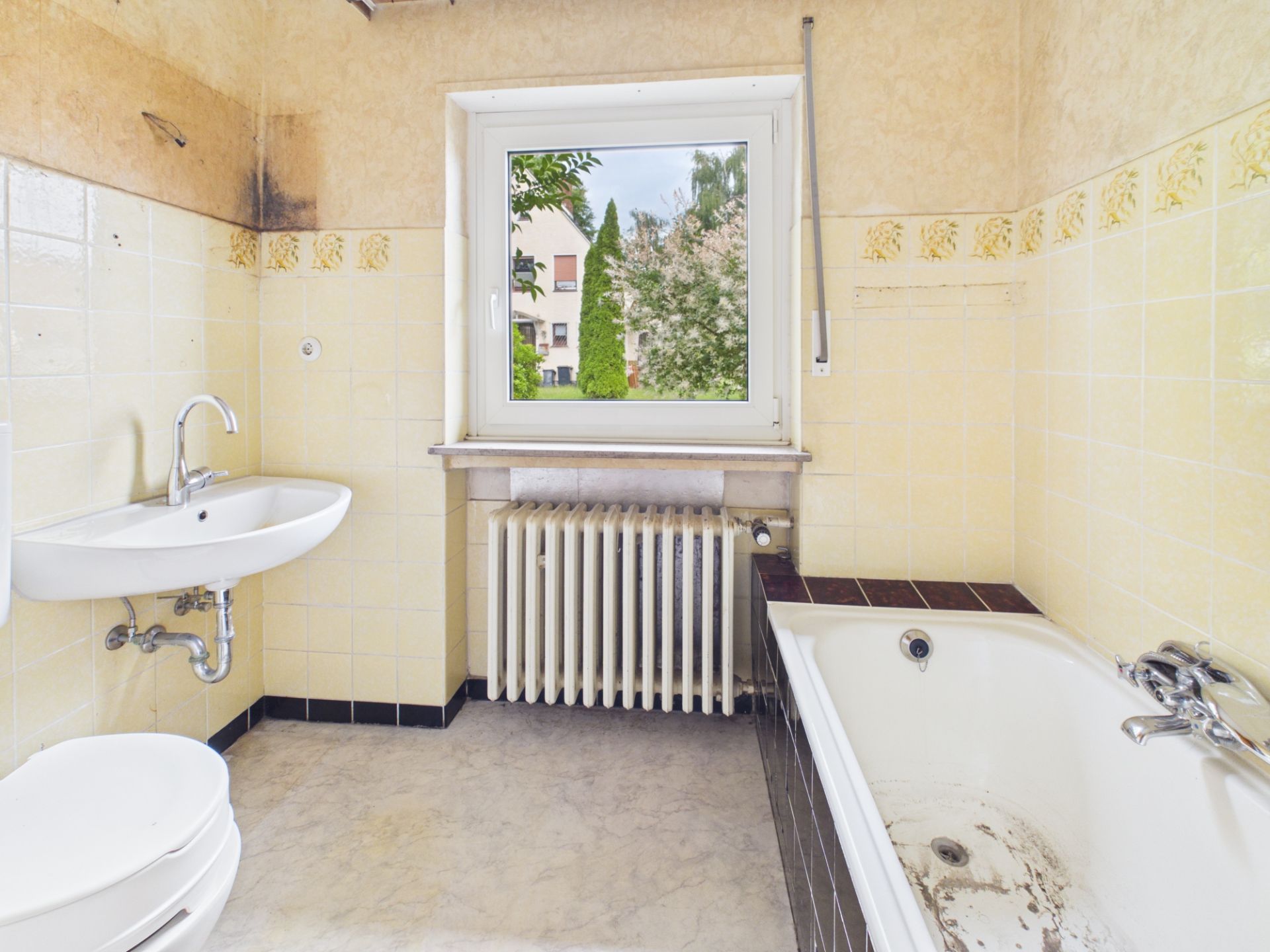 Altes Badezimmer mit Badewanne, Waschbecken, Toilette und Fenster mit Ausblick.
