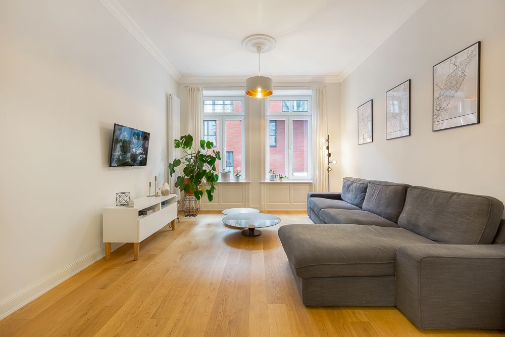Helles Wohnzimmer mit grauem Sofa, Holzboden, Couchtisch, Fernseher und großen Fenstern.