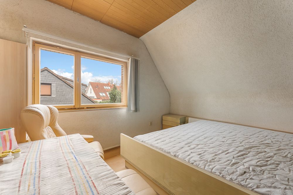 Helles Schlafzimmer mit großem Fenster, Bett, Schreibtisch und Sessel unter schräger Decke.