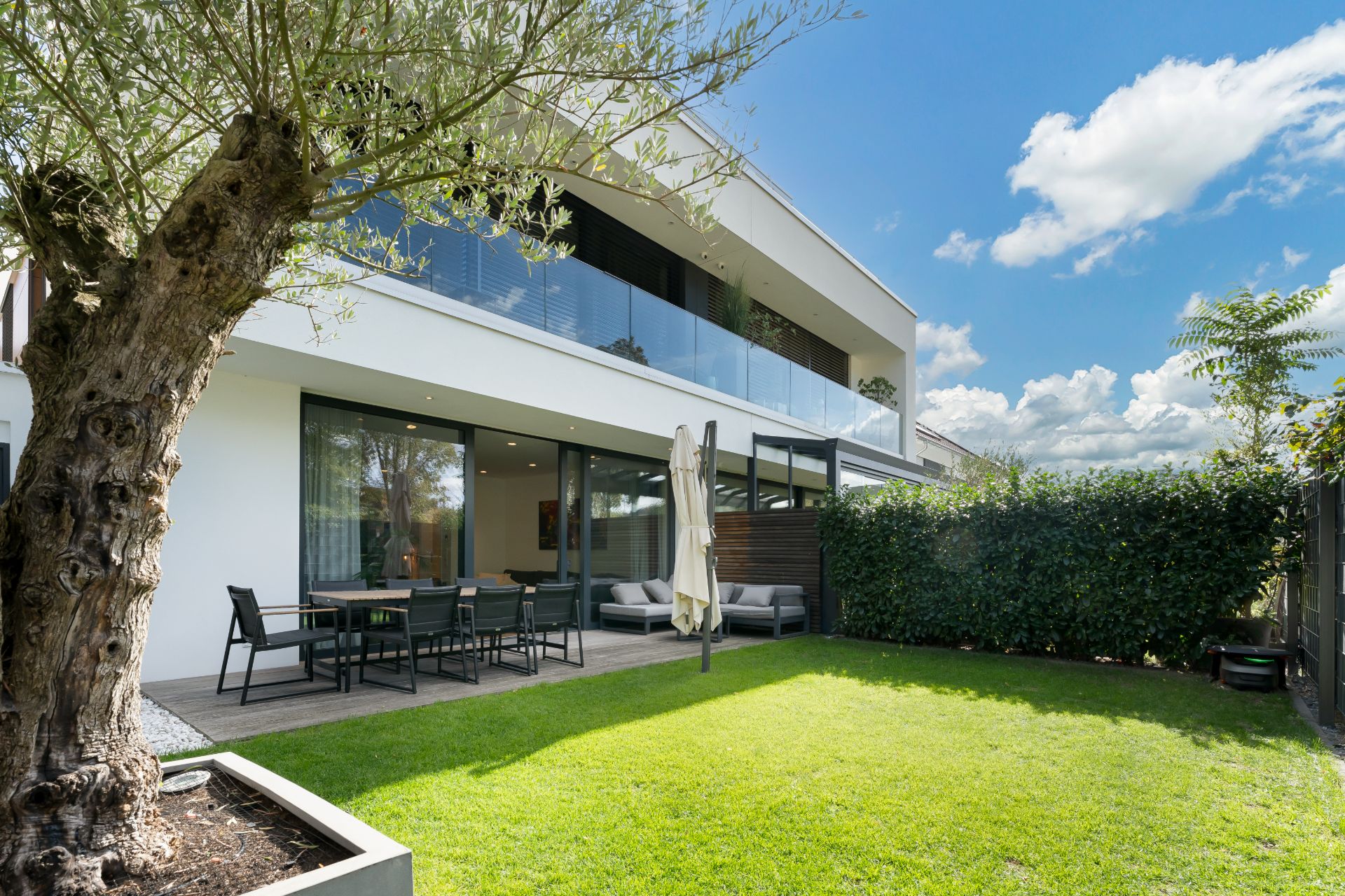 Moderner Garten mit Terrasse, Esstisch, Stühlen, Sofa und Baum vor einem Wohnhaus.