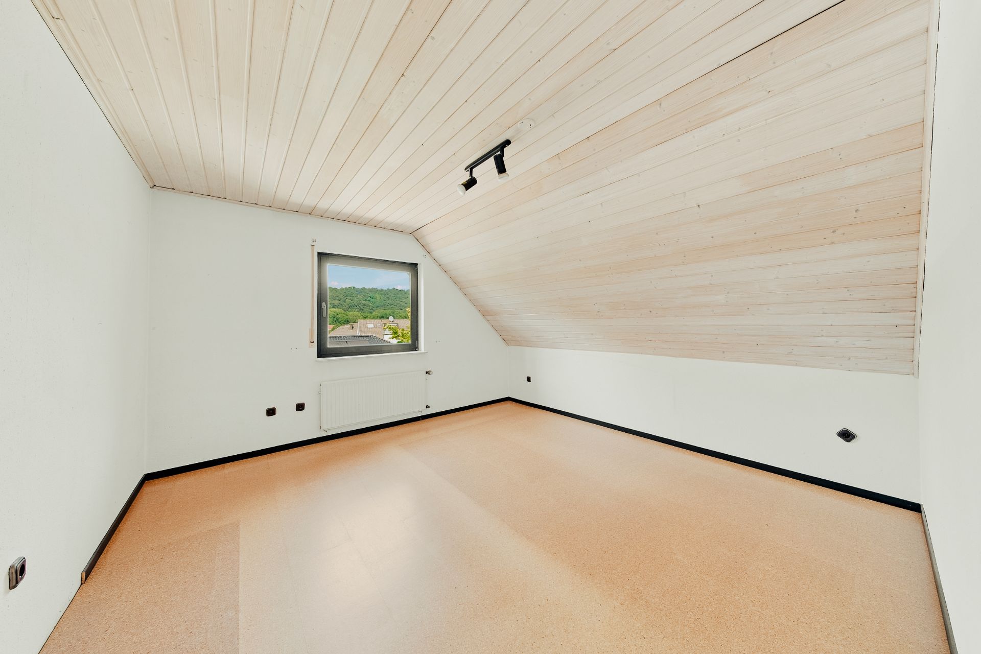 Leeres Zimmer mit schräger Holzdecke, Fenster und heller Bodenfläche.