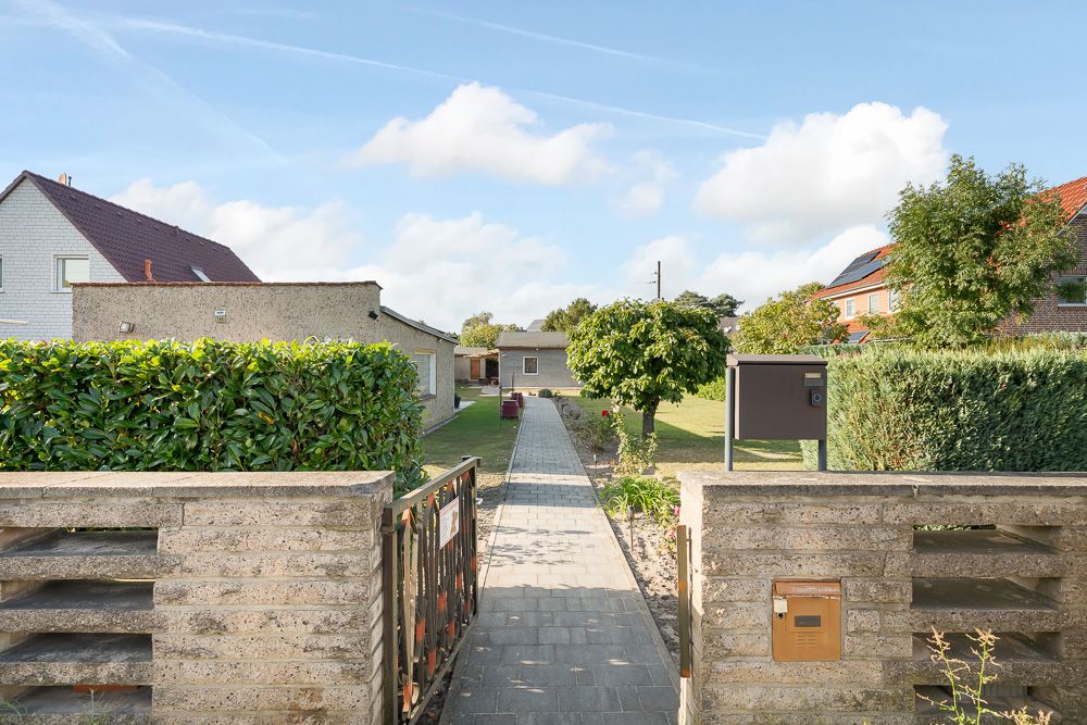 Eingangstor mit gepflastertem Weg zu einem Haus, umgeben von Garten und Bäumen.