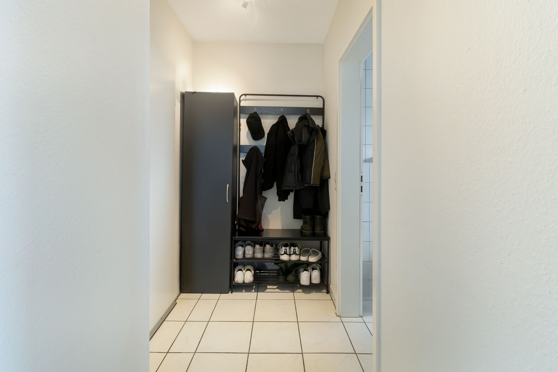 Flur mit Garderobe, Schuhregal und grauem Schrank auf weiß gefliestem Boden.