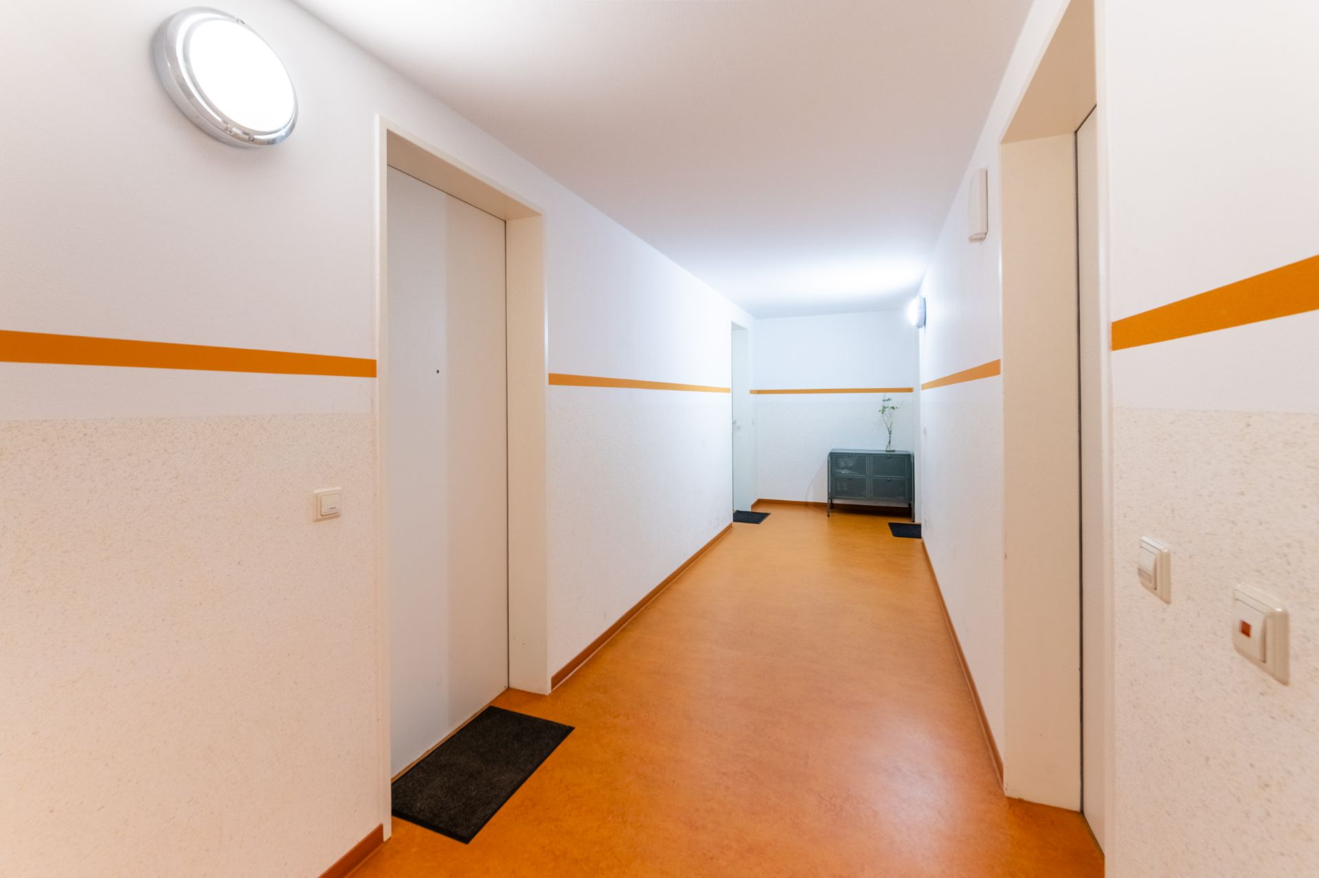 Heller Flur mit orangefarbenem Boden und Türen, Wandleuchten und einer Kommode am Ende.
