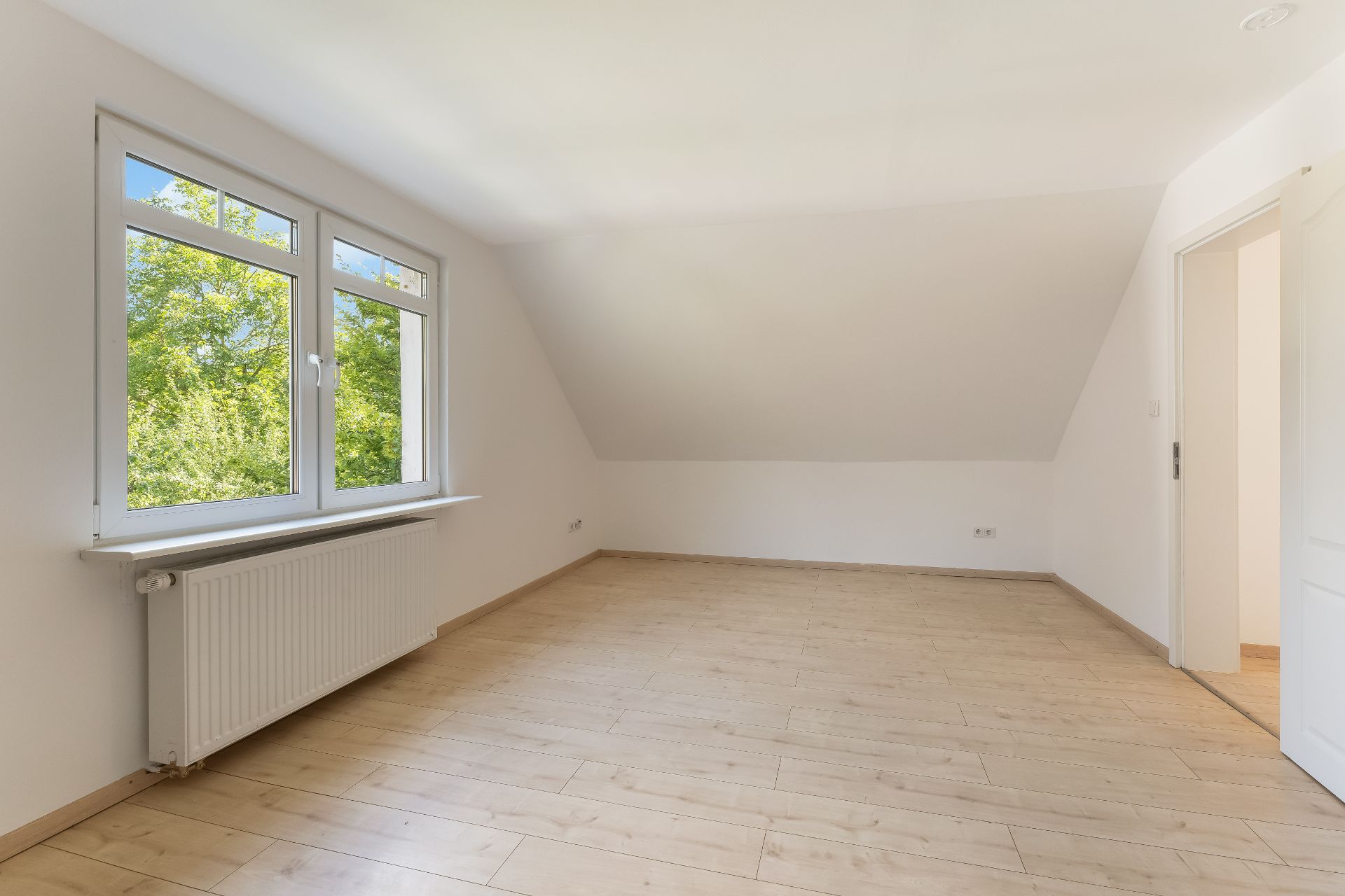 Leeres Zimmer mit Holzfußboden, großem Fenster und Heizung unter dem Fenster.