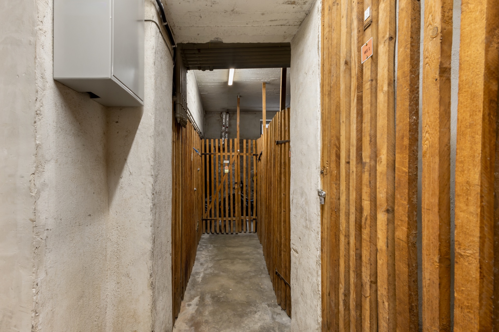 Schmaler Kellerflur mit Holzgittertüren und Betonwänden.