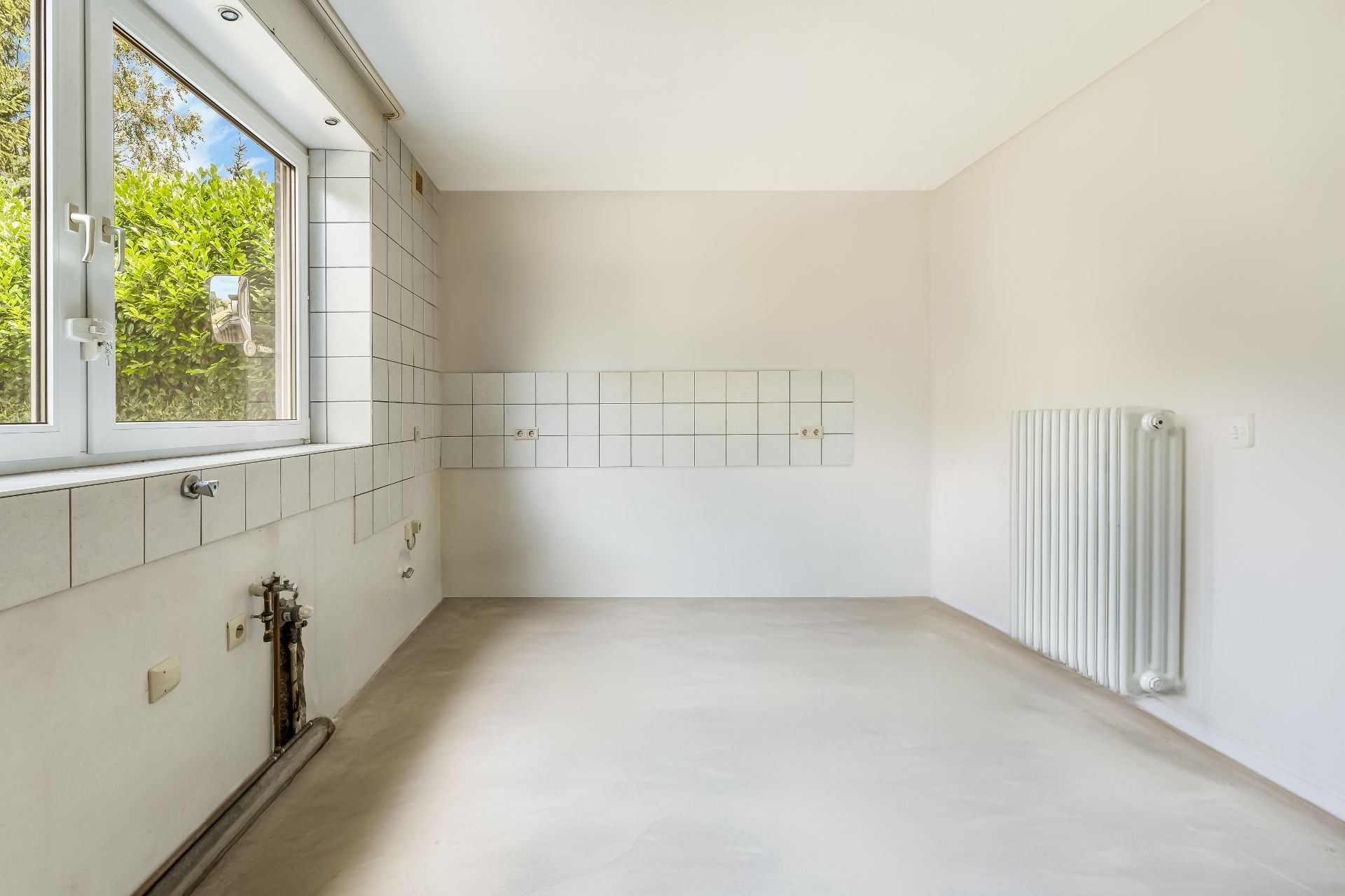 Leerer, heller Raum mit großem Fenster, weißen Fliesen und Heizkörper an der Wand.