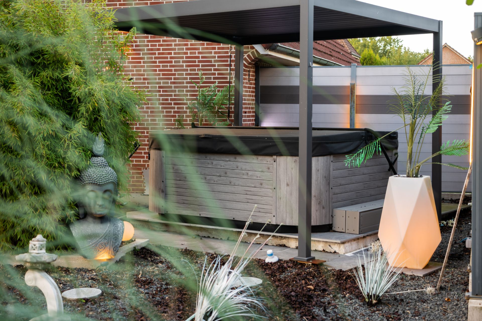 Garten mit Whirlpool unter Pergola, Buddha-Statue, Pflanzen und moderner Beleuchtung.