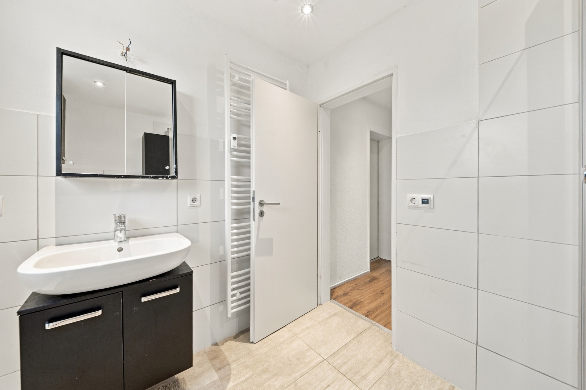 Modernes Badezimmer mit Waschbecken, Spiegel, Heizkörper und geöffneter Tür zum Flur.
