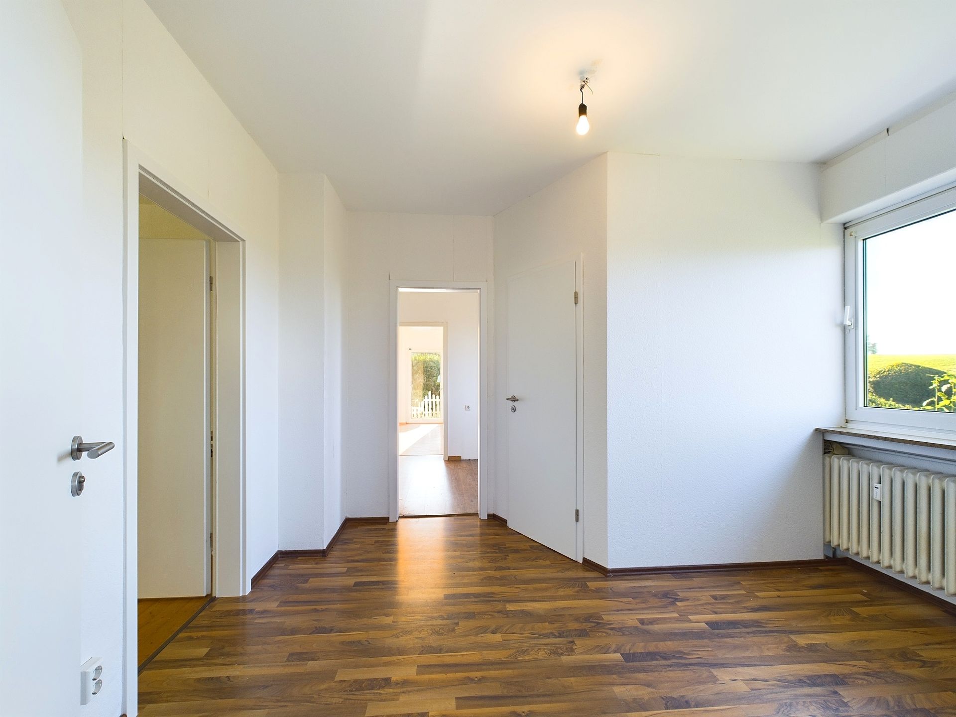 Leerer Raum mit Holzboden, weißen Wänden, Fenster und mehreren Türen.