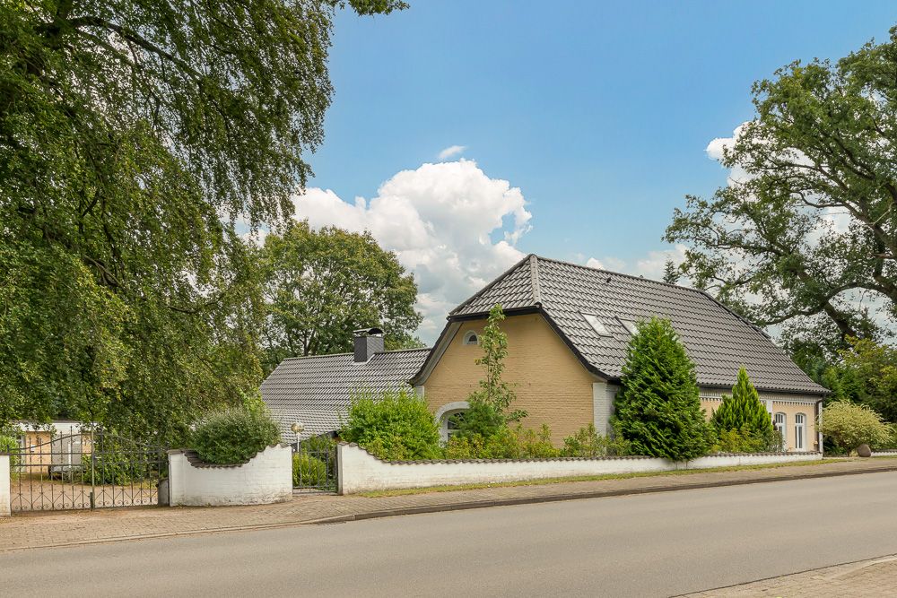 Haus mit grauem Dach, gelber Fassade und Garten hinter einer weißen Mauer an einer Straße.
