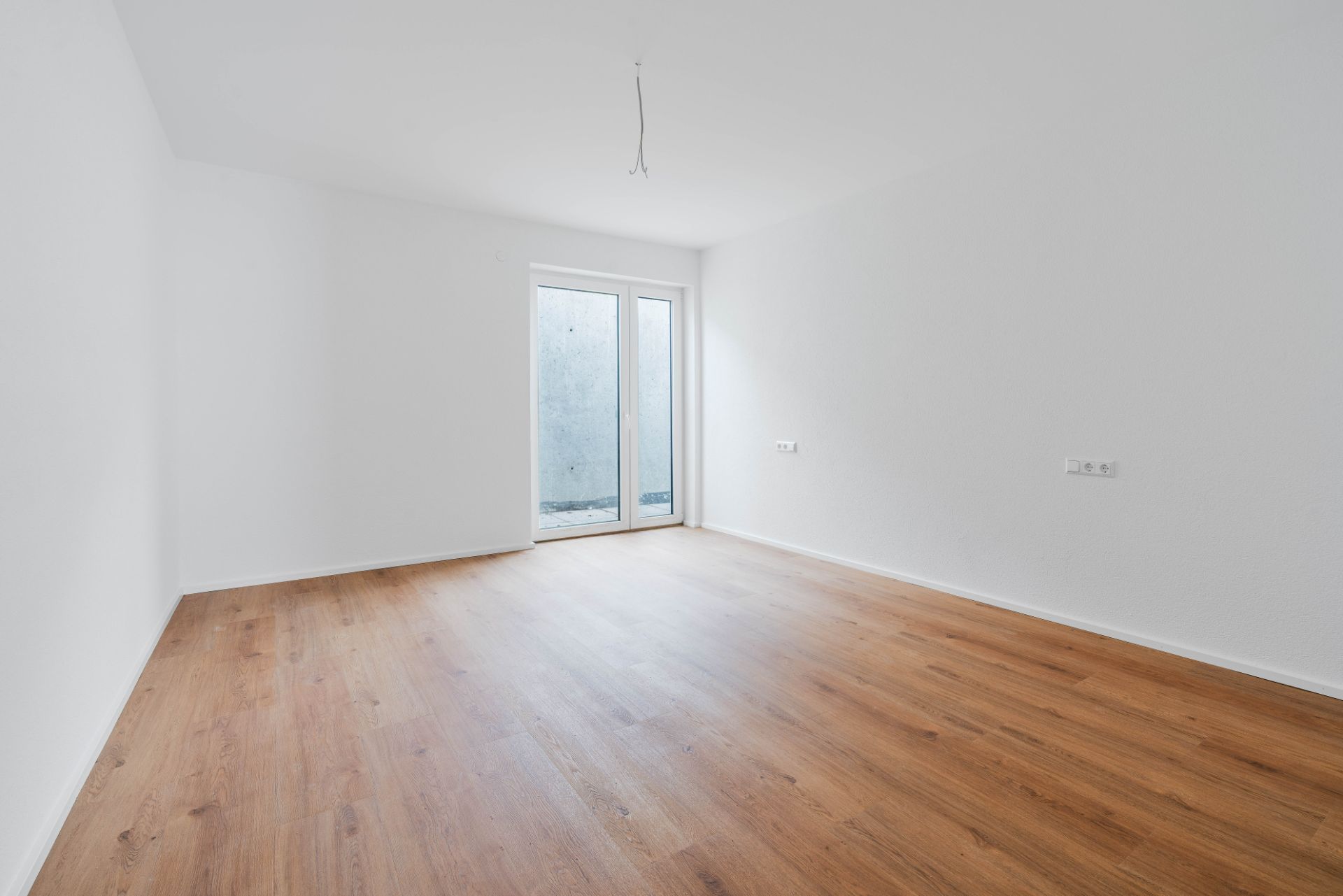 Leerer Raum mit hellem Holzboden, weißen Wänden und Glastür zu einer Terrasse.