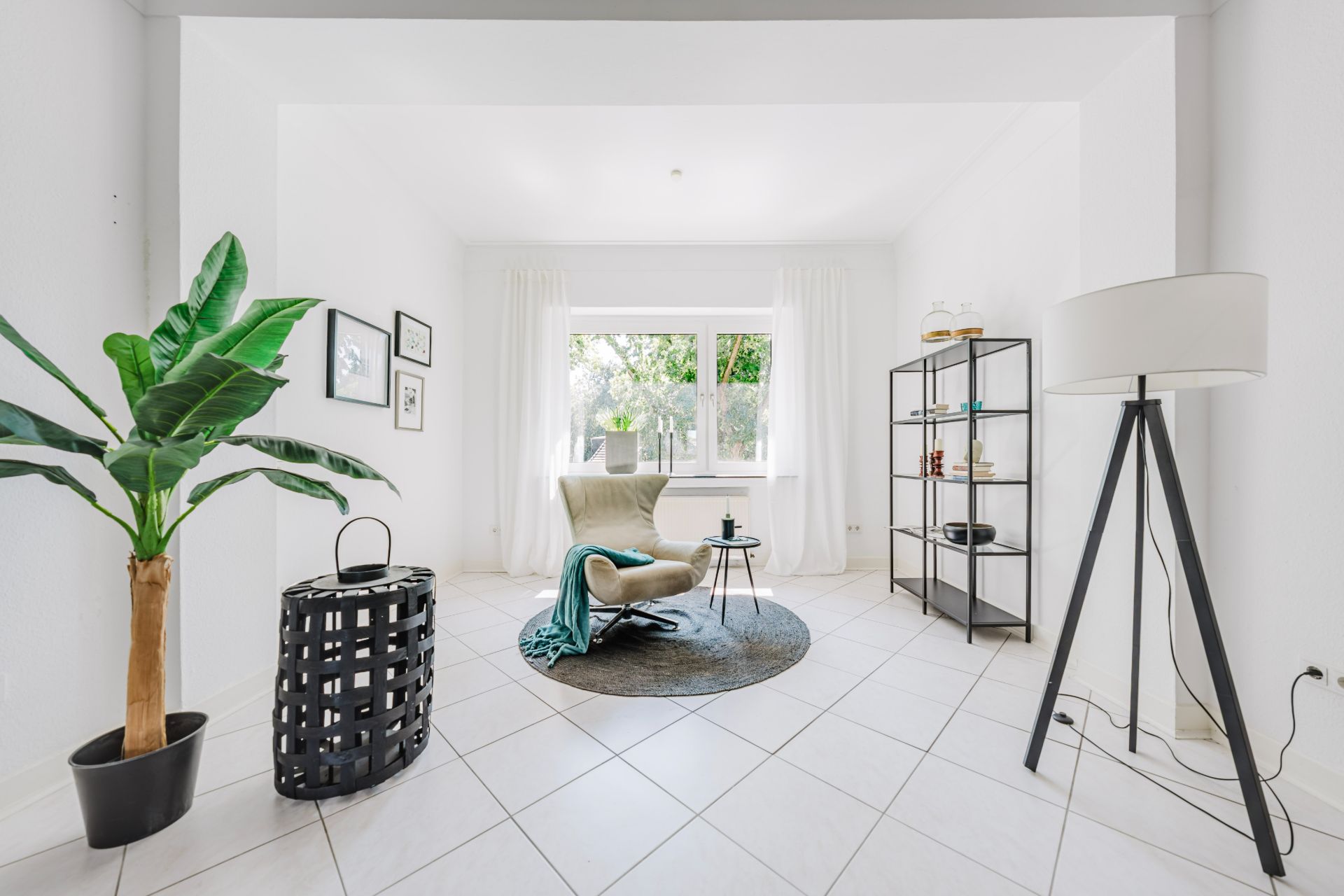 Helles Wohnzimmer mit Sessel, Regal, Stehlampe, Pflanze und großem Fenster.
