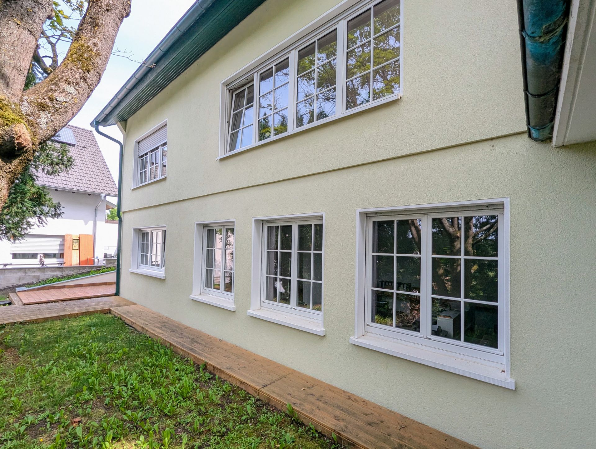 Heller Außenbereich mit mehreren weißen Fenstern und grünem Garten vor dem Haus.