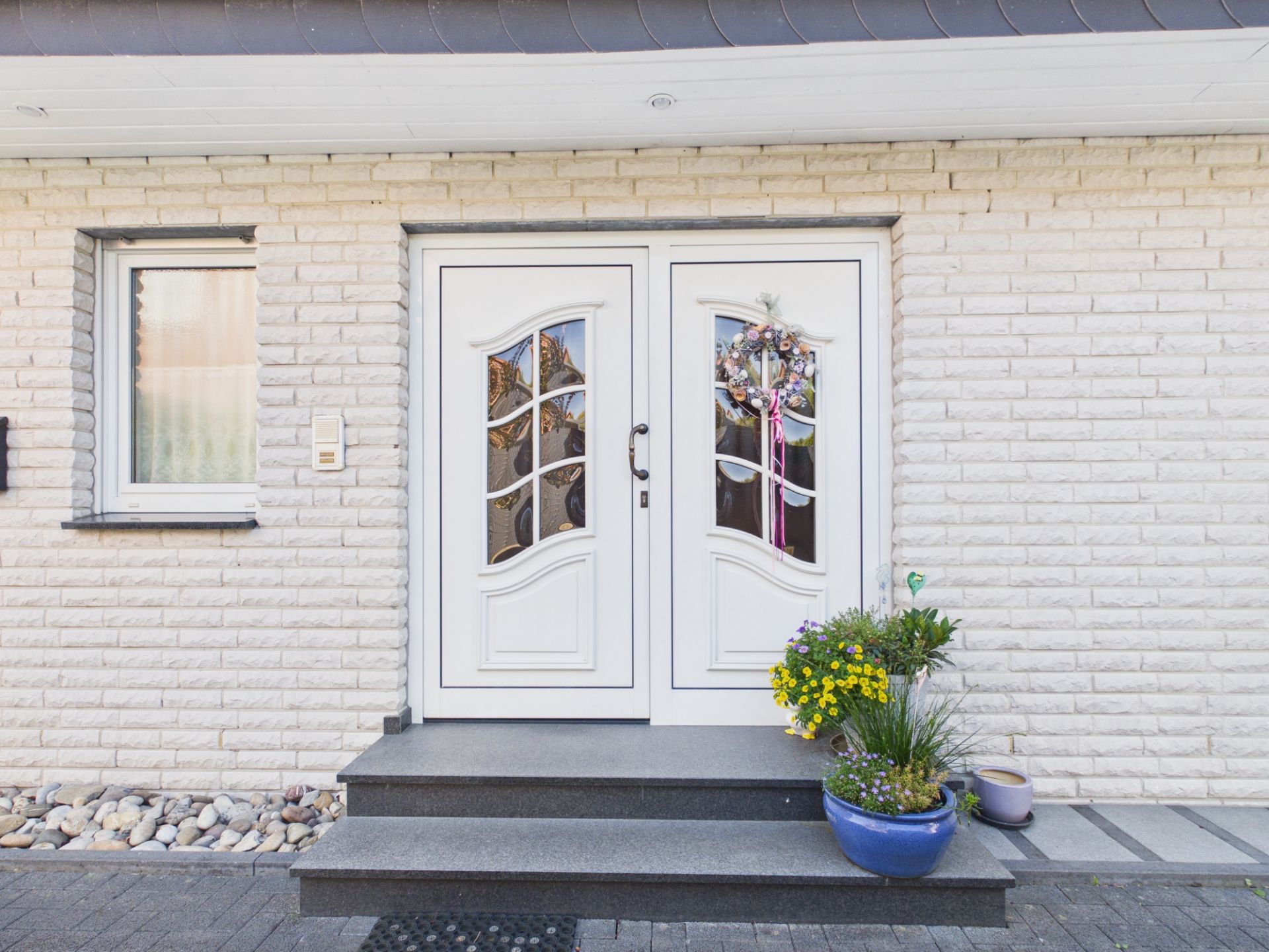 Weiße Haustür mit zwei Flügeln, Blumenschmuck und Topfpflanzen auf der Treppe.