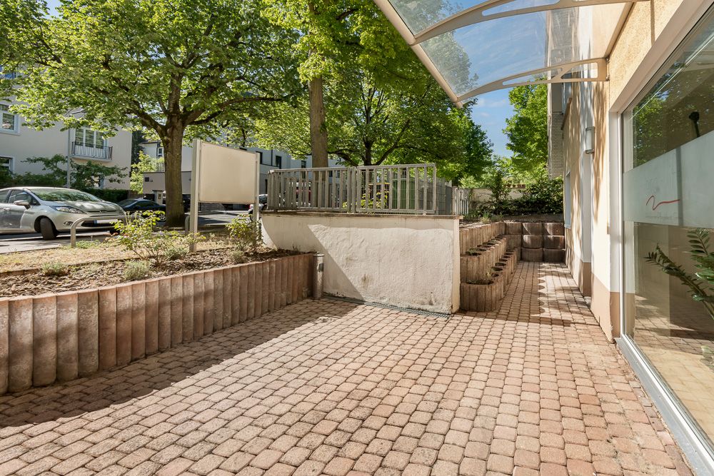 Gepflasterte Terrasse mit Hochbeeten und Glasüberdachung neben Wohnhaus, Bäume im Hintergrund.