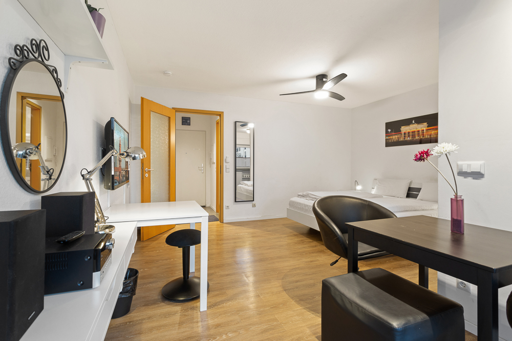 Helles Wohnstudio mit Bett, Schreibtisch, runder Spiegel, Esstisch und Holzboden.