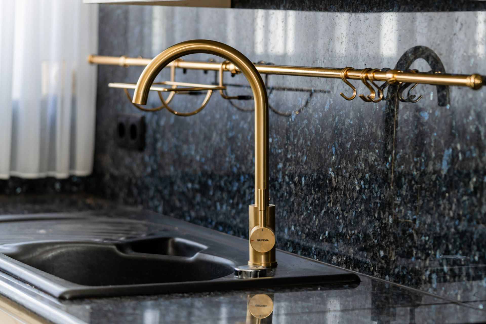 Goldener Wasserhahn und schwarze Spüle vor einer dunklen, glänzenden Küchenrückwand.