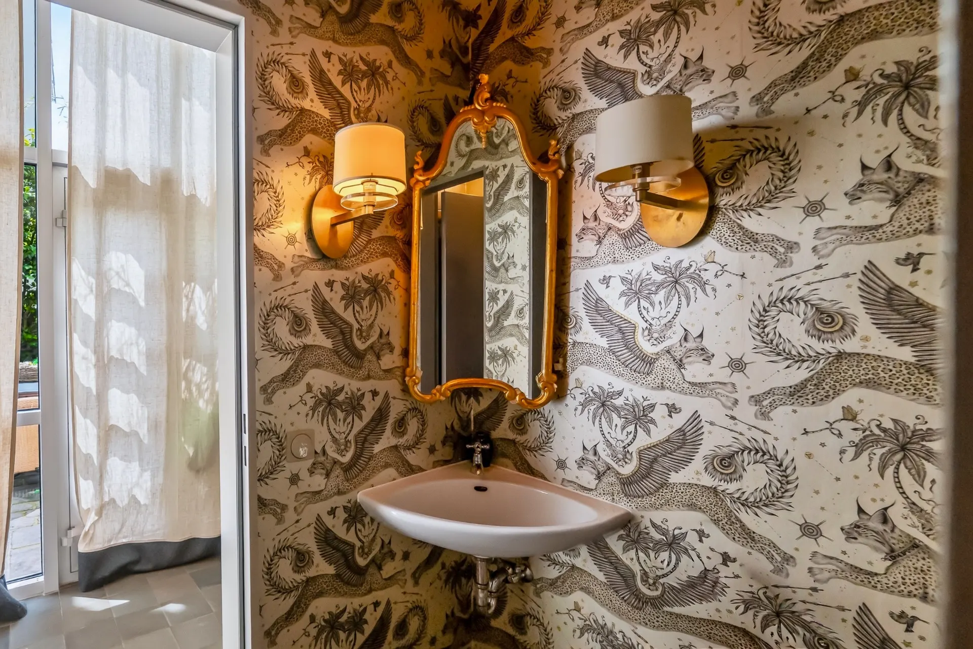 Kleines Badezimmer mit Wandwaschbecken, Spiegel, zwei Lampen und gemusterter Tapete mit Tiermotiven.