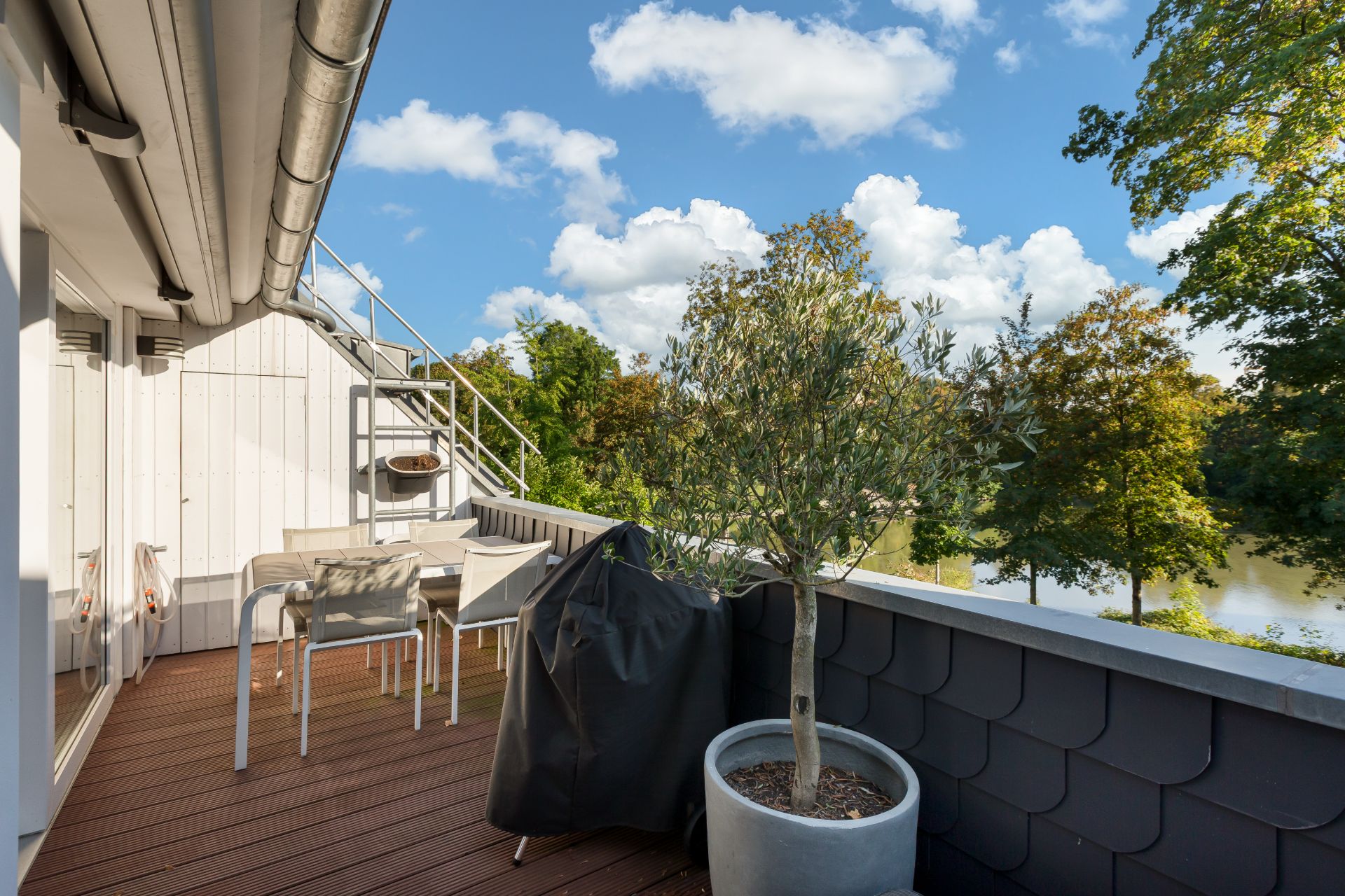 Balkon mit Holzboden, Sitzgruppe, Grillabdeckung und großem Pflanzkübel, Blick auf Bäume.