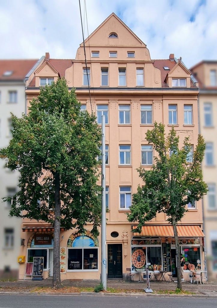 Fünfstöckiges Wohnhaus mit Geschäften im Erdgeschoss, zwei Bäumen und Gehweg davor.