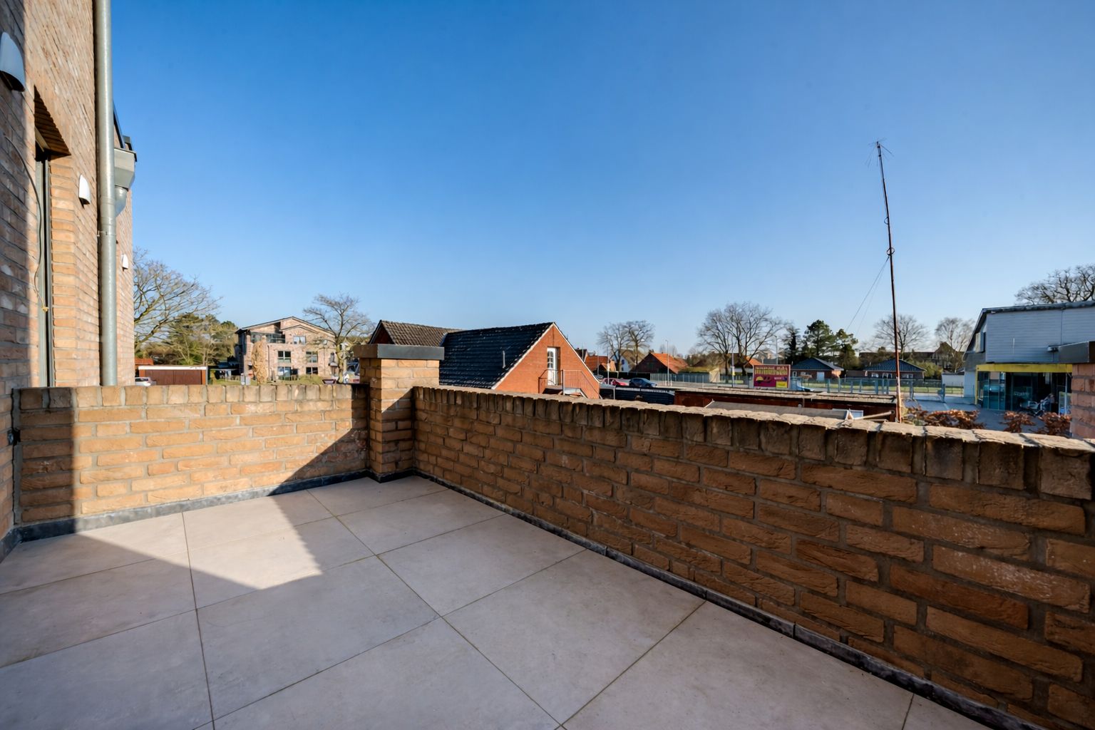 Große Terrasse mit Fliesenboden und Backsteinmauer, Blick auf Nachbarhäuser und blauen Himmel.