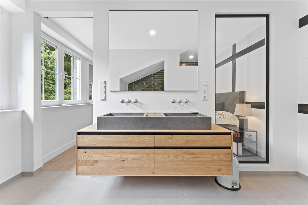 Modernes Bad mit Holzwaschtisch, großem Spiegel und grauem Waschbecken. Fenster links, Schlafzimmer sichtbar rechts.