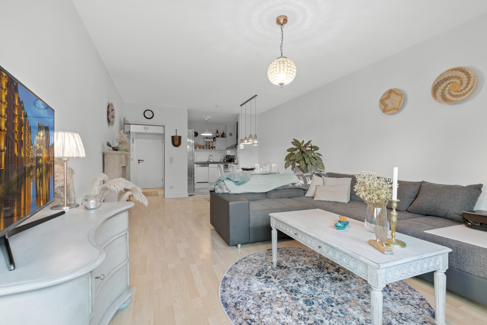 Helles Wohnzimmer mit Ecksofa, Couchtisch, Fernseher und offener Küche im Hintergrund.