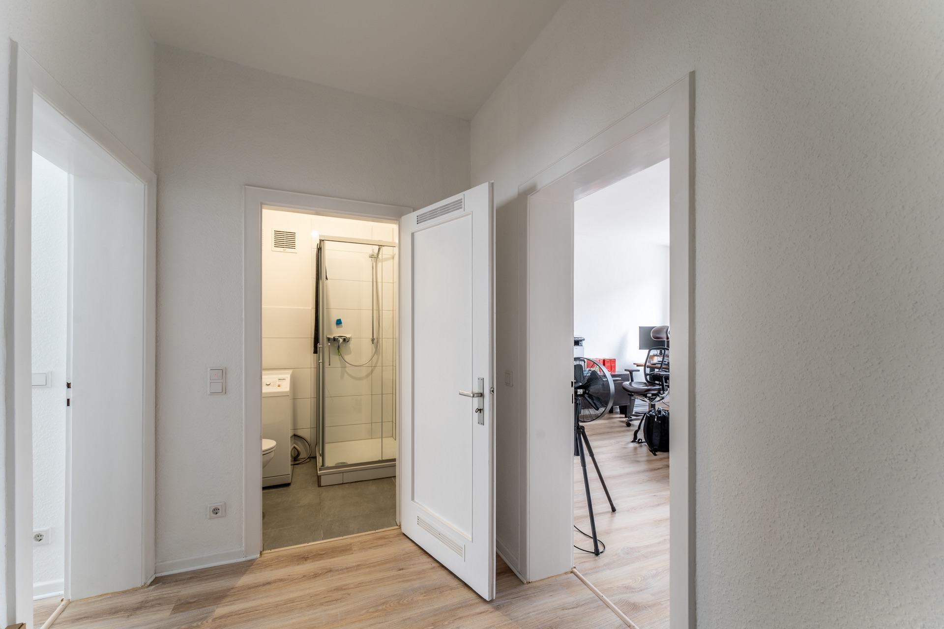 Flur mit Blick in ein Bad mit Dusche und ein Arbeitszimmer mit Stuhl und Monitor.