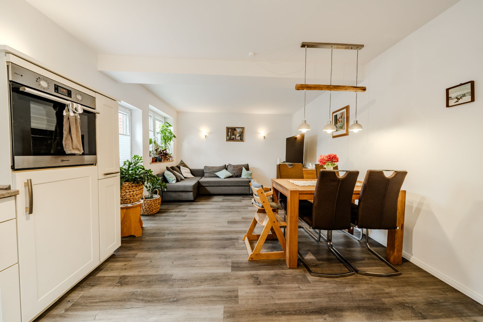 Helles Wohnzimmer mit Ecksofa, Esstisch, Stühlen und offener Küche mit Einbauofen.