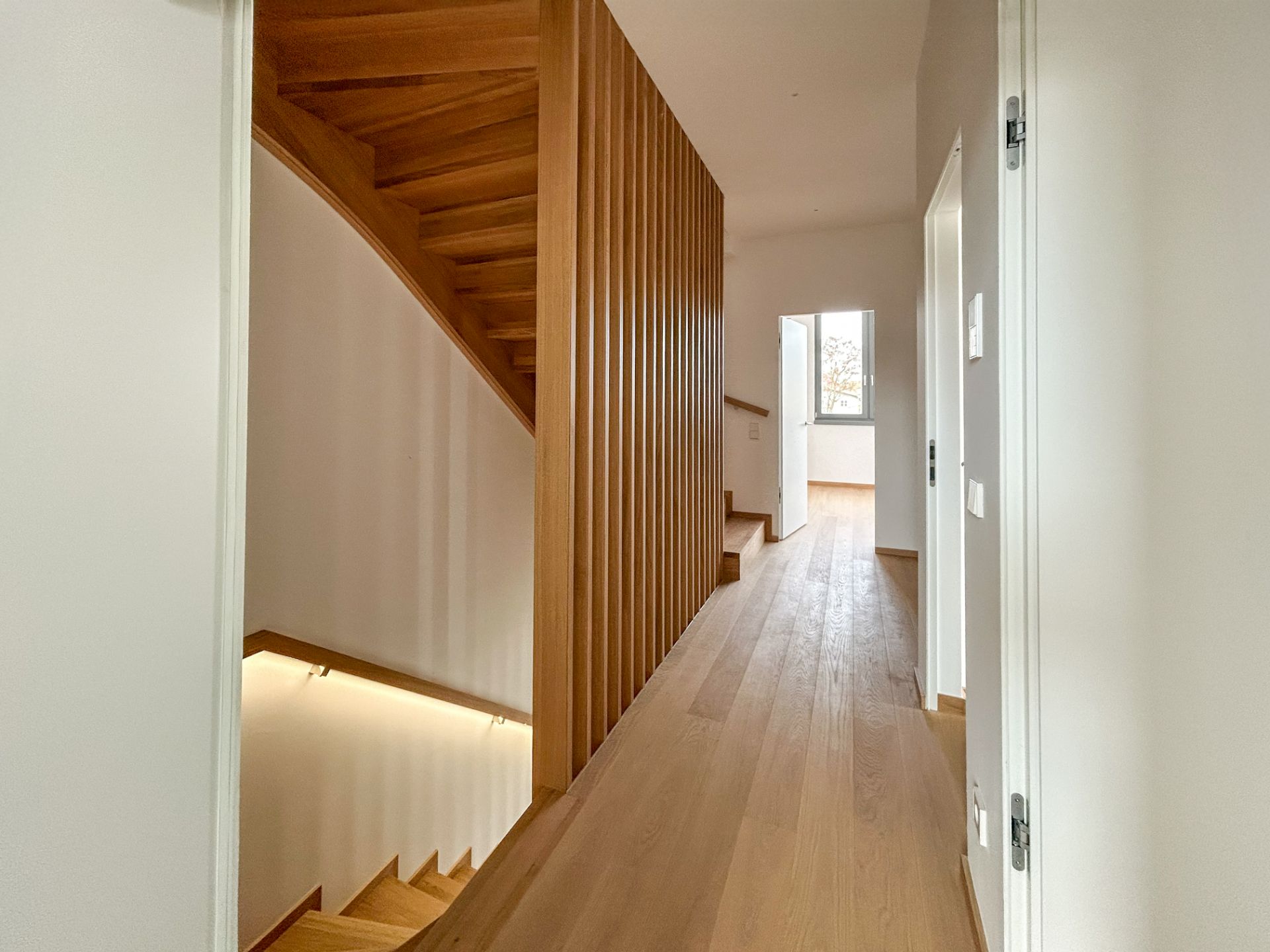 Heller Flur mit Holztreppe und Holzgeländer, Türen und Fenster am Ende des Gangs.