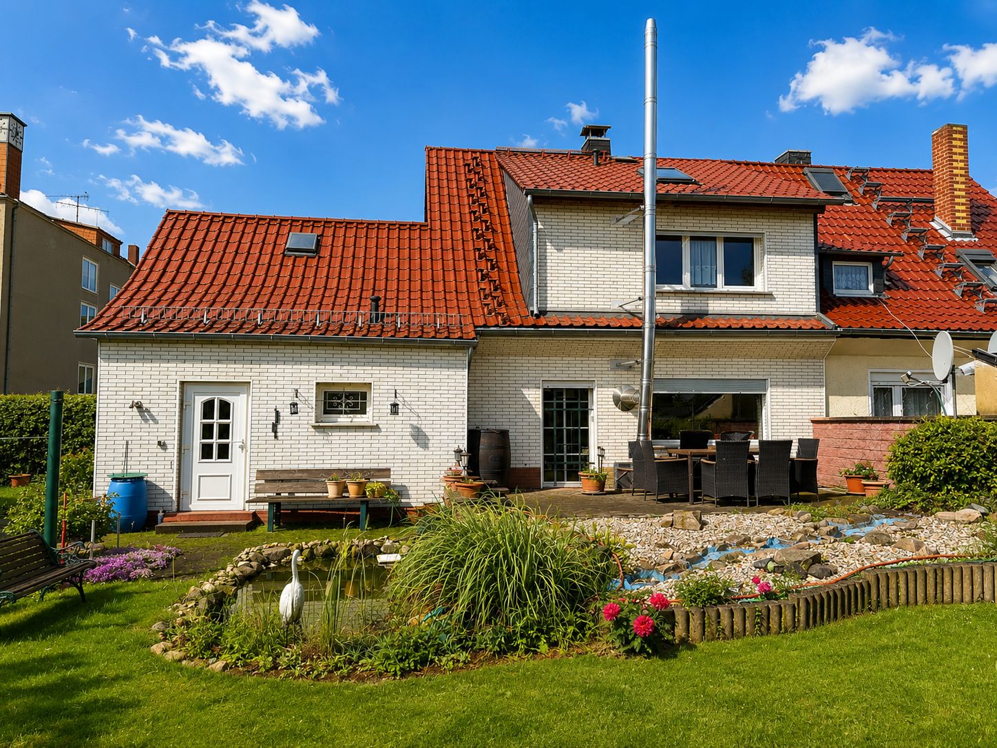 Rückseite eines Hauses mit rotem Dach, Terrasse, Garten und kleinem Teich im Vordergrund.