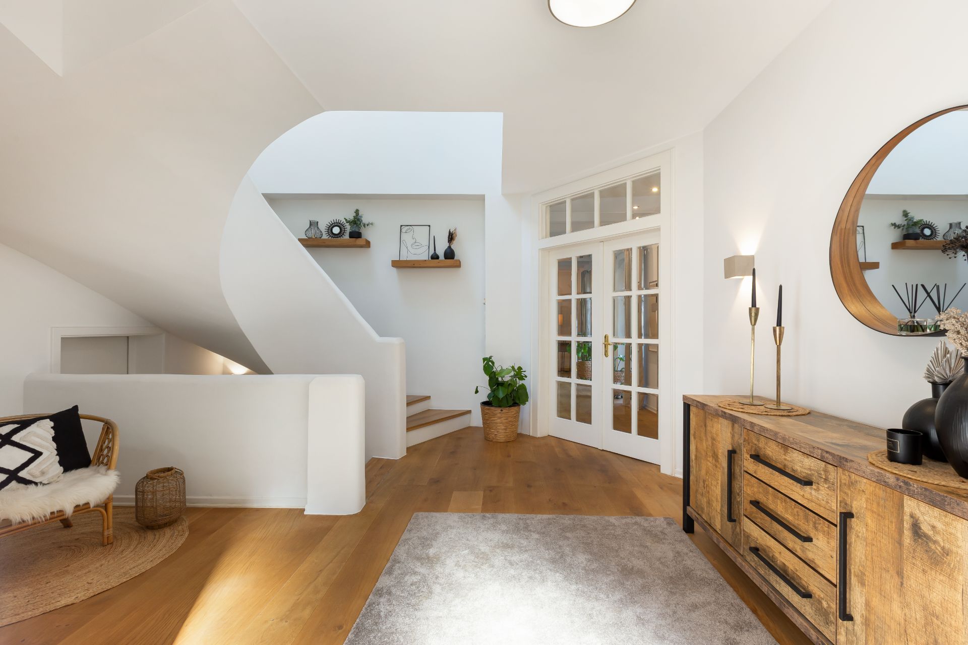 Heller Flur mit Holzkommode, rundem Spiegel, Treppe und großen Glastüren.