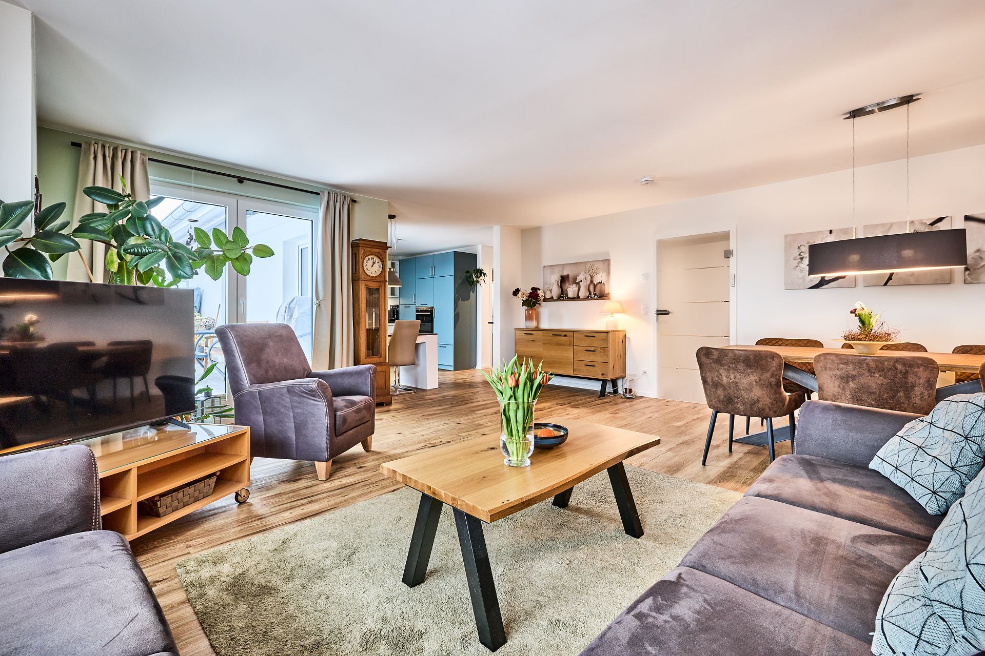 Modernes Wohnzimmer mit Sofa, Sesseln, Fernseher, Esstisch und großer Pflanze.