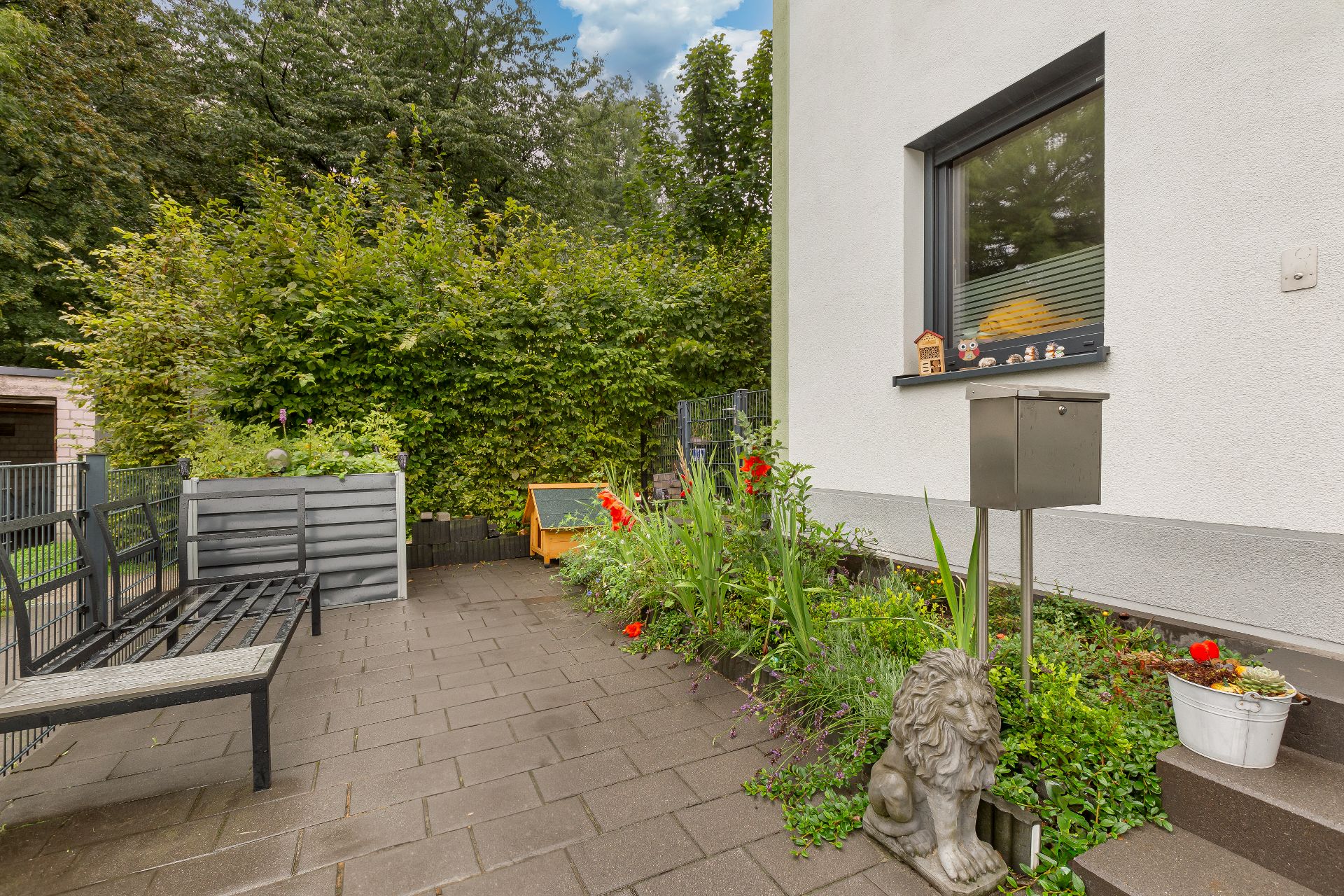 Gepflasterte Terrasse mit Bank, Blumenbeet, Löwenstatue und Briefkasten vor einem Haus.