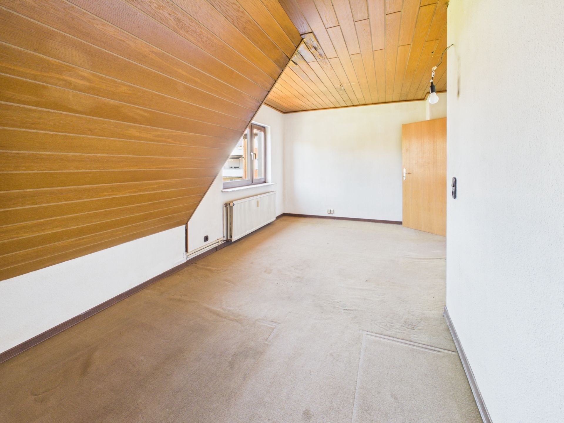 Leeres Zimmer mit schräger Holzdecke, Fenster und braunem Teppichboden.