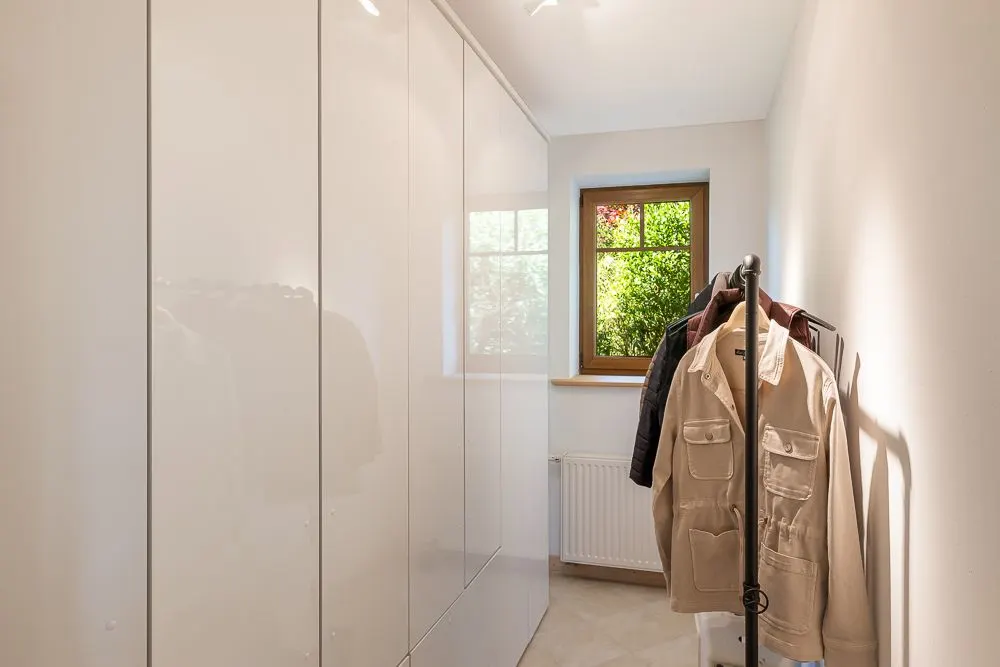 Heller Ankleideraum mit weißem Schrank, Fenster und Kleiderständer mit Jacken.