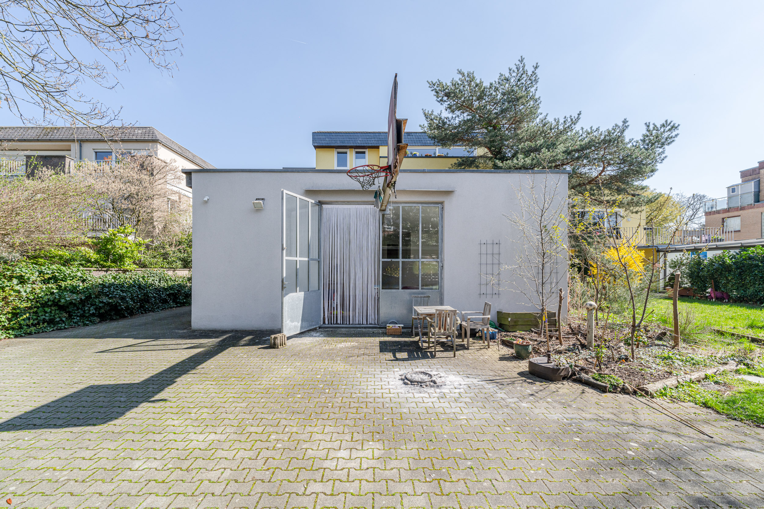 Kleines weißes Gebäude mit Basketballkorb, Terrasse und Gartenmöbeln im Garten.