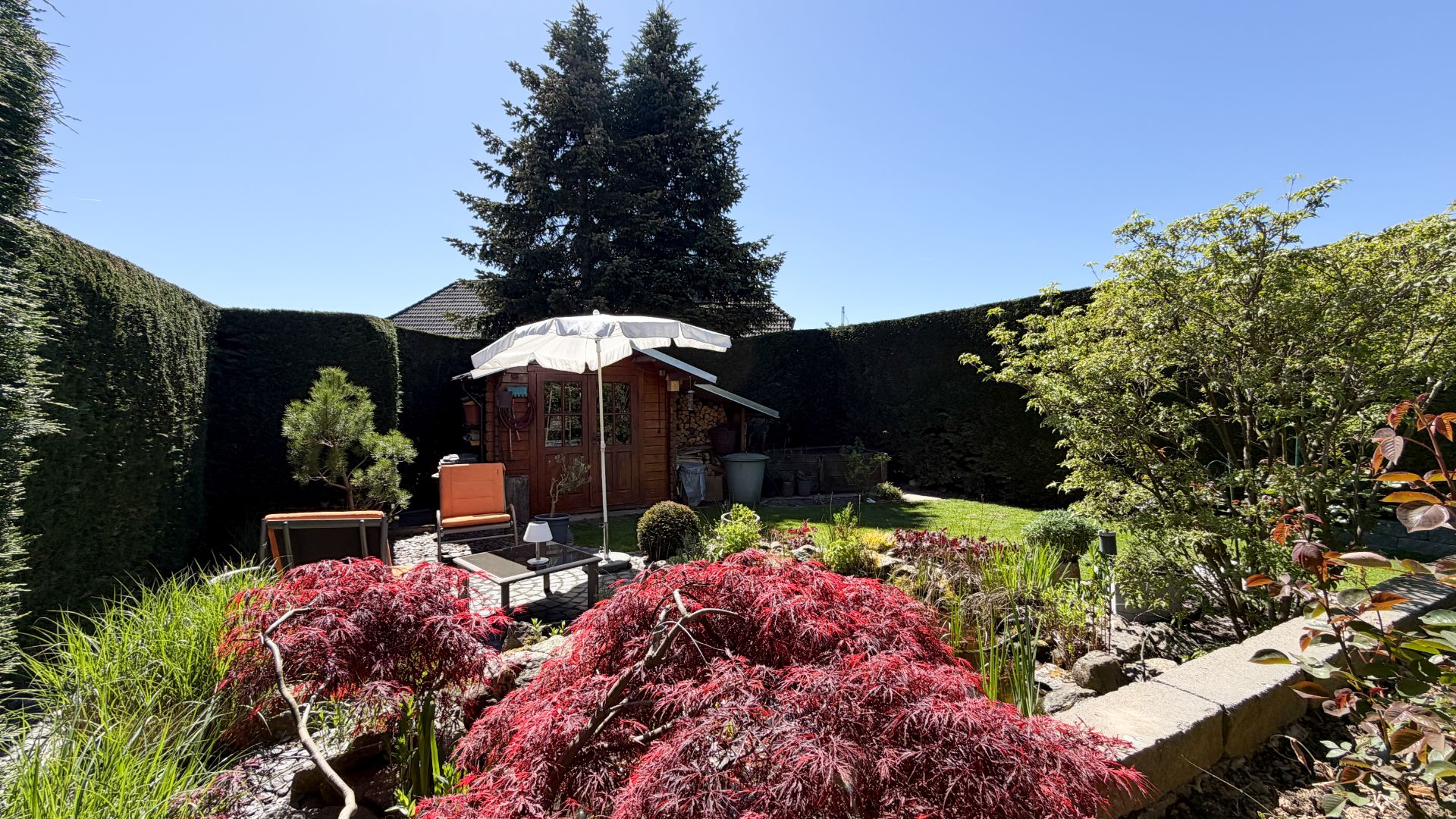 Garten mit Holzhaus, Sitzbereich mit Sonnenschirm und Teich, umgeben von Hecken.