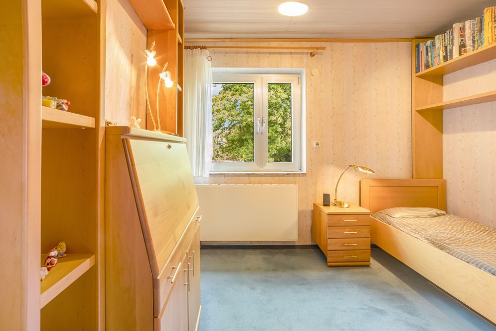 Helles Schlafzimmer mit Einzelbett, Schreibtisch, Regal und Fenster mit Blick ins Grüne.